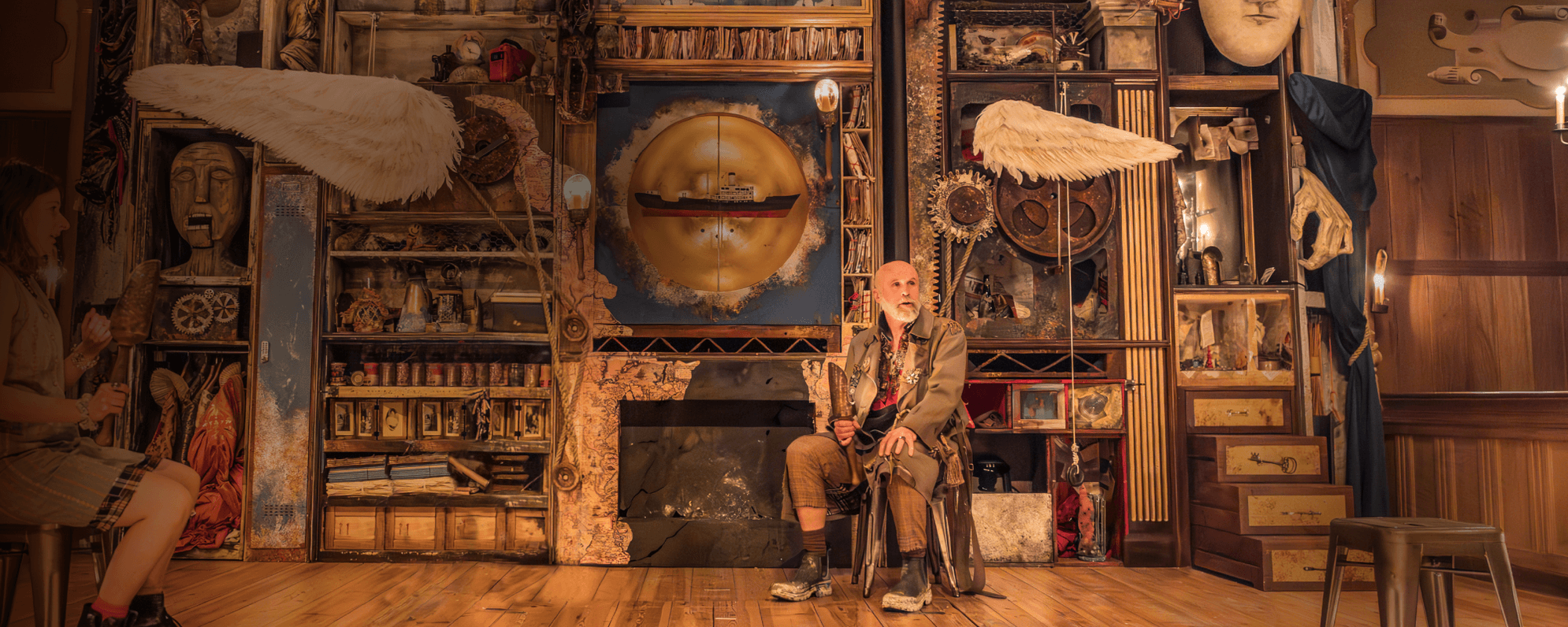 The Tempest. A person sits on a wooden stage holding a golden candle sconce. They are wearing a long khaki jacket and muddy black ankle boots. The wall behind them is covered in various objects: maps, cogs, ropes, books, angel wings and a model of a boat.