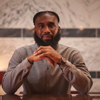 Daniel Bailey, who wears a grey shirt, looking directly at the camera over folded hands