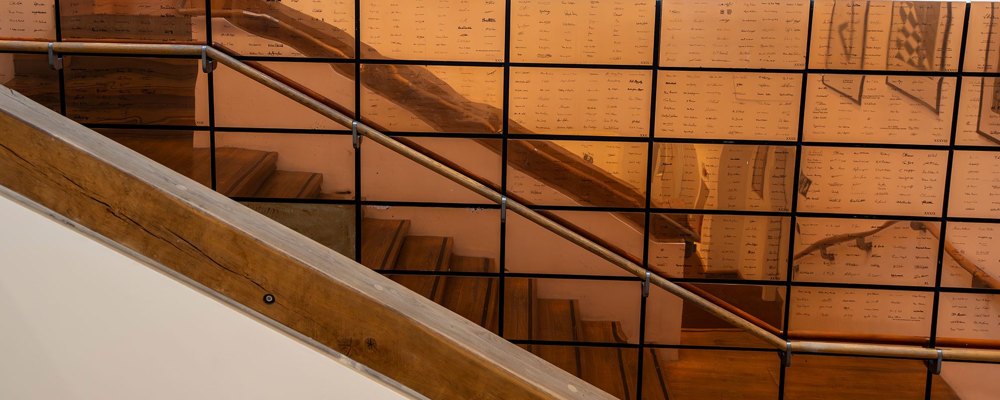 The side view of a wooden staircase. Behind it, bronze squares cover the wall.
