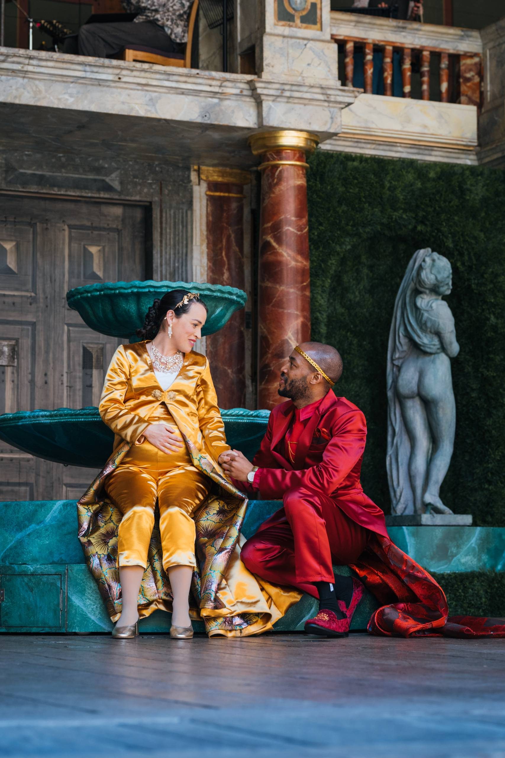 A pregnant woman with dark hair, wearing a yellow satin suit and gown, sits on the edge of a green fountain. Crouching next to her and holding her hand is a man wearing a red satin suit and gown.