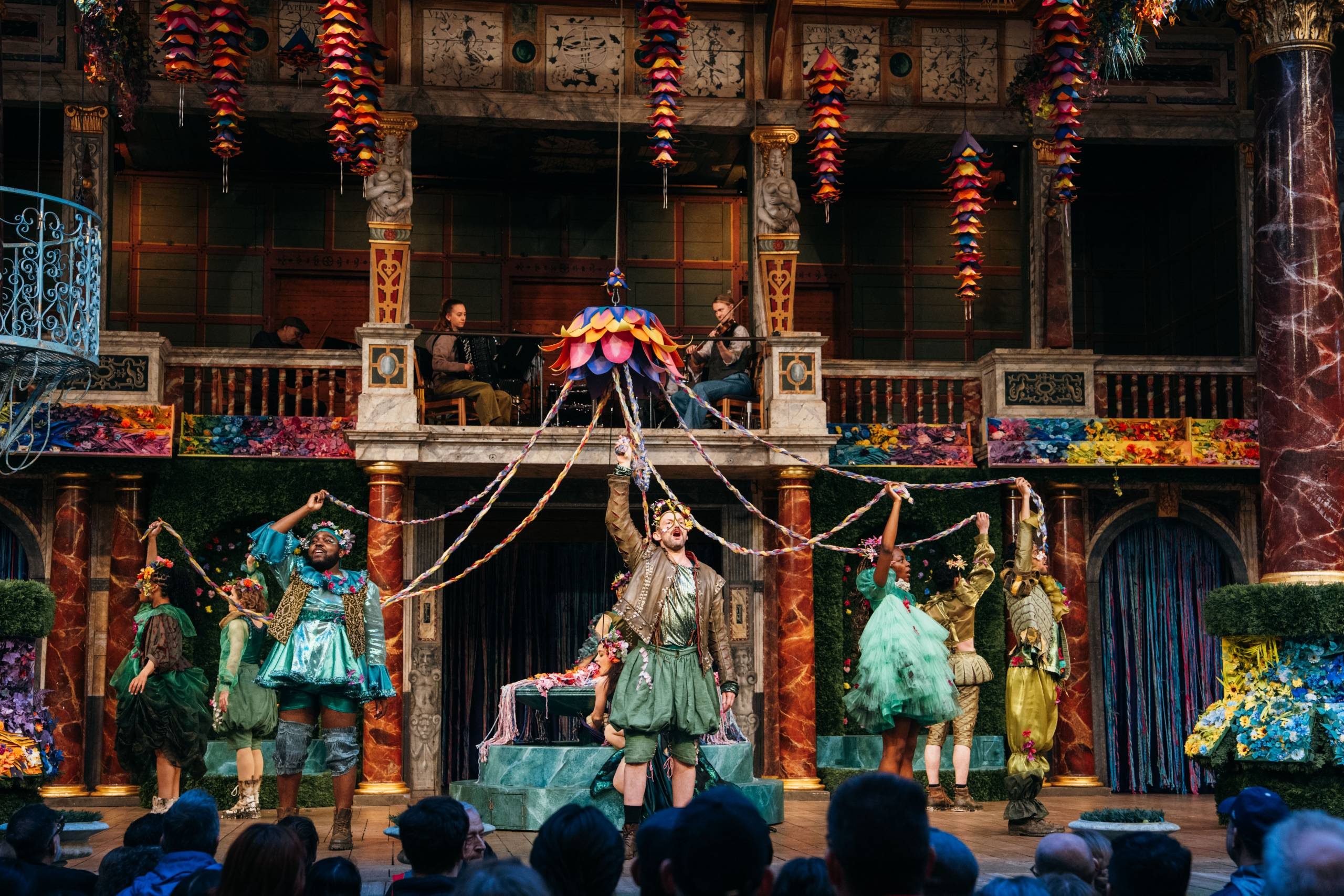 People wearing green outfits, decorated with flowers, stand in a circle holding coloured strands that lead off of a colourful decoration hung mid-air.