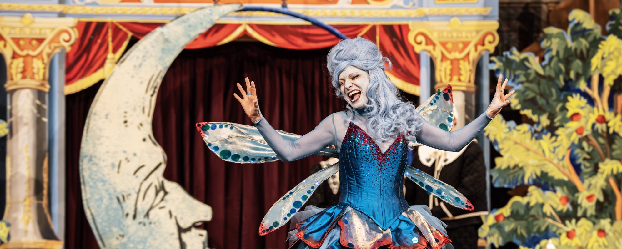 Lucy McCormick as Blue Fairy stands on stage in front of a wooden crescent moon. She wears a light blue wig and a blue frilly dress with a red trim and blue wings attached.