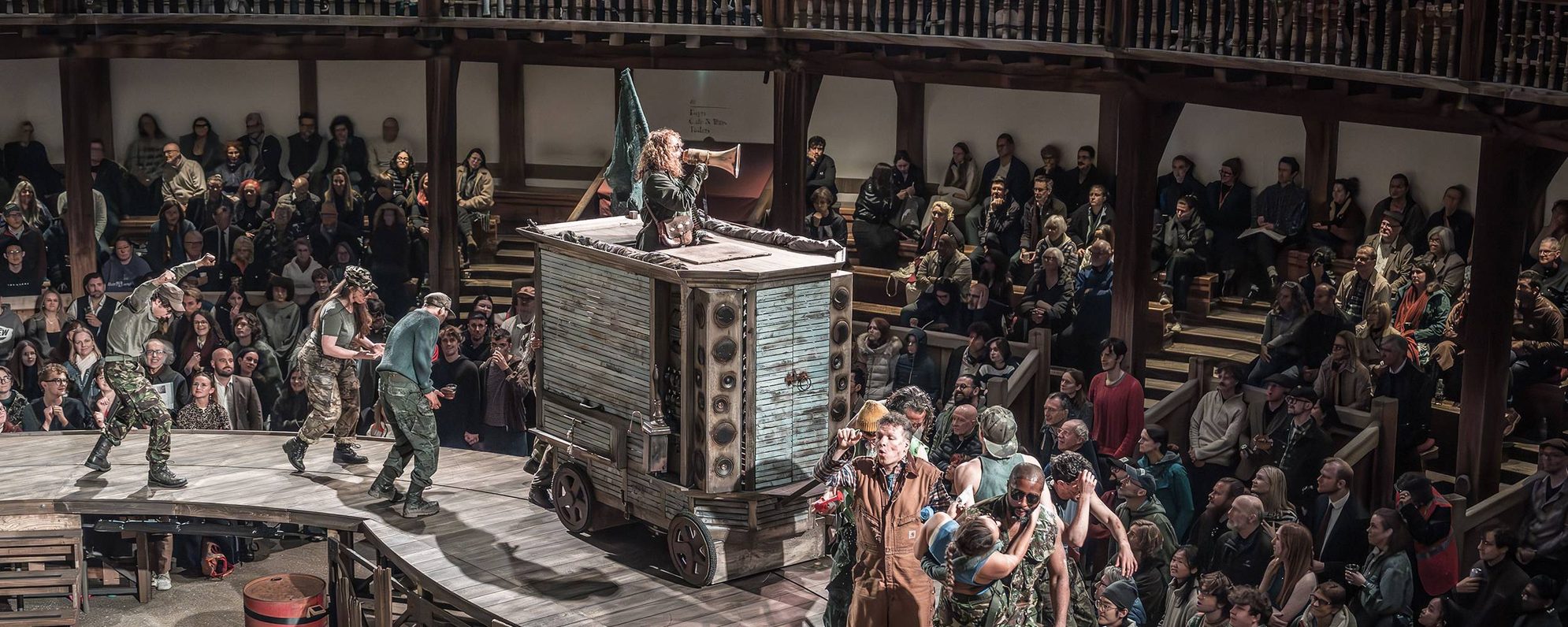 In a round theatre with wooden seating galleries, an audience watches as a parade of people wearing camouflage dance around a large blue cart. Standing in the cart is a white woman with long curly hair, speaking into a megaphone.