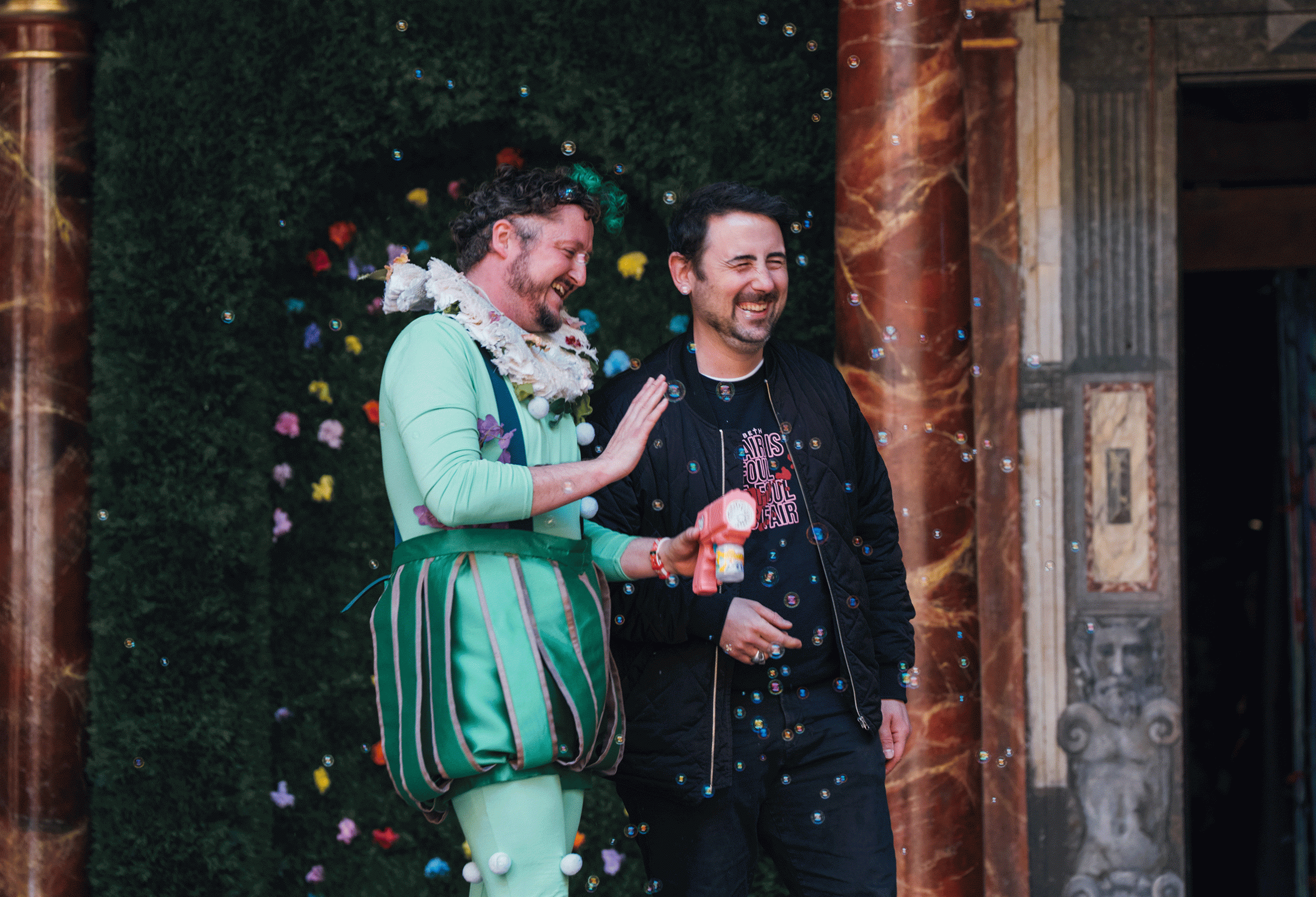 A man wearing a green Elizabethan costume and holding a pink bubble gun stands beside a person wearing a black jacket and trousers. Both are smiling. There are bubbles in the air around them.