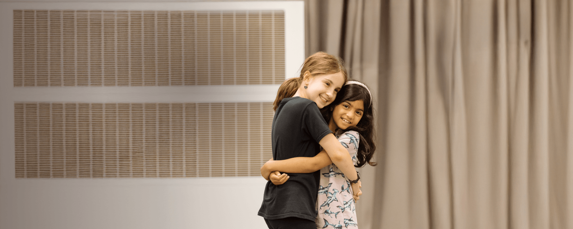 Two children stand in a room, hugging and smiling.