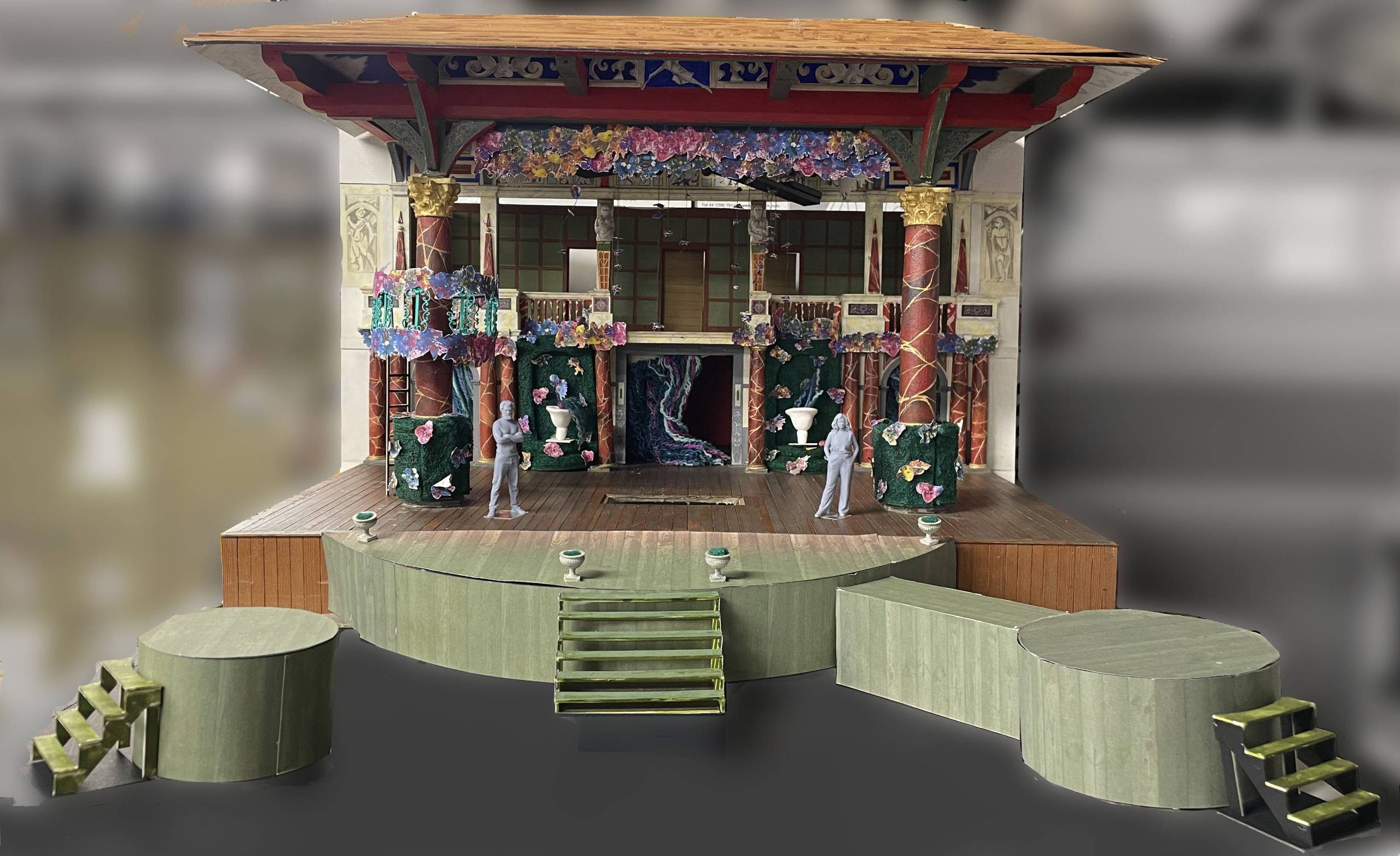 A model of a wooden theatre with a wooden roof held up by red marble pillars, a green circular jetty with green steps and a round green platform with green steps.