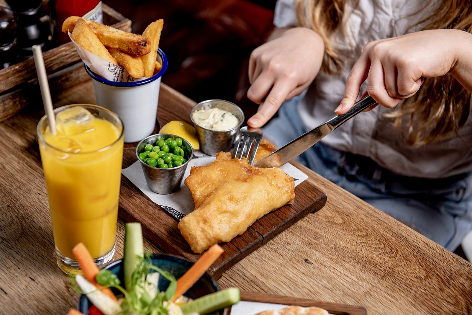 A person is cutting into a piece of battered fish with a knife and fork. Next to the fish on the table are peas, sauces, chips, cucumber and carrot sticks and a glass of orange juice.