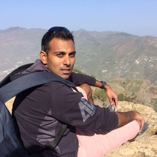 Professor Pablo Mukherjee sitting on a hill and looking back at the camera. Behind him is a view of clifftops in the distance.