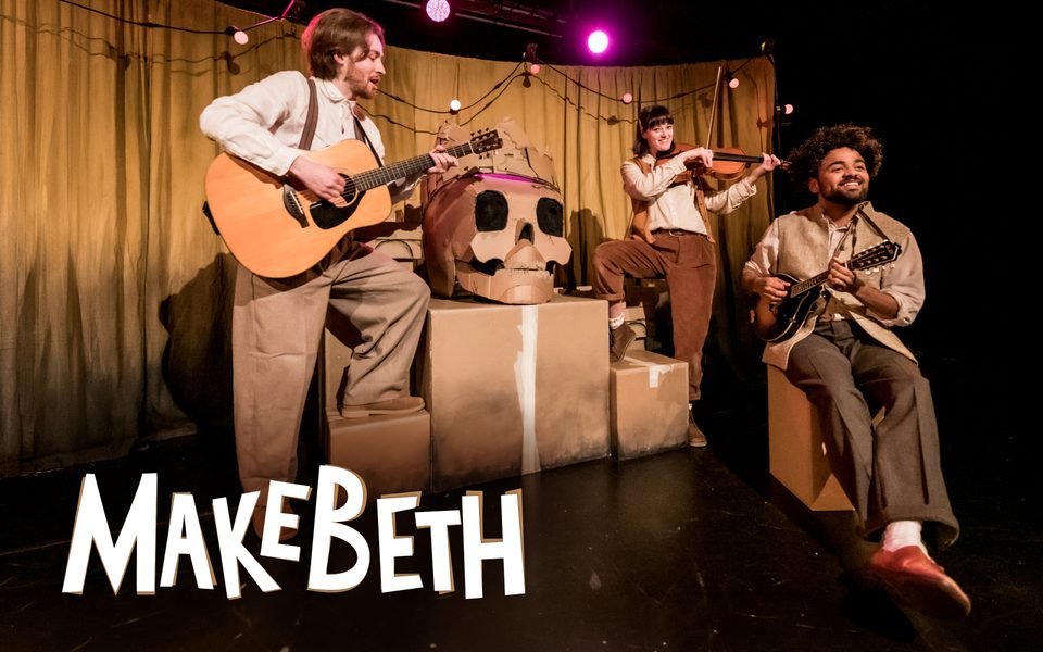 Makebeth. Three musicians, wearing brown and beige, play two guitars and a violin as they gather around cardboard boxes. On top of the boxes sits a giant cardboard skull with a crown.