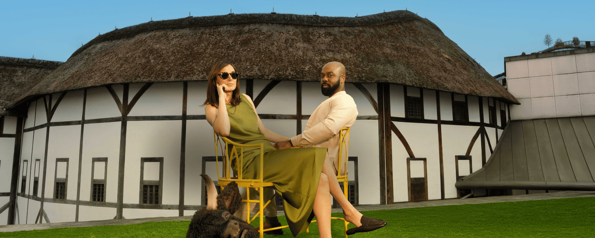 Pippa Nixon, a white woman with shoulder-length brown hair, and Ken Nwosu, a bald man with brown skin and a beard, are seated on a bench on grass outside a white building with a thatched roof. There is a realistic cow mask beside them.