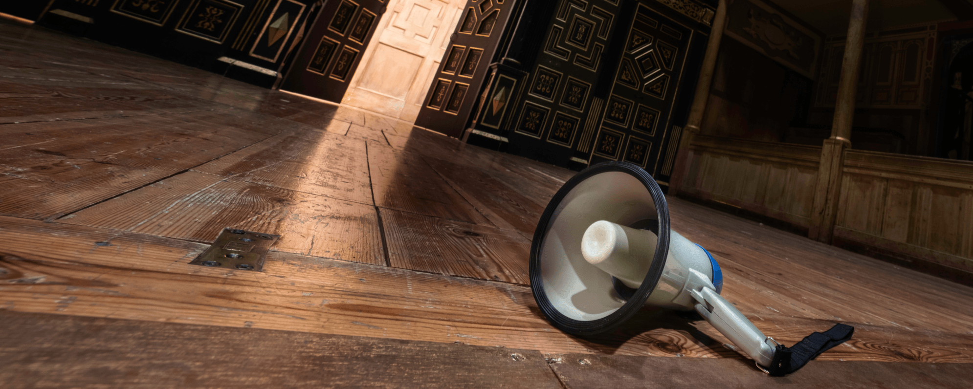 A white and blue megaphone lies on a wooden stage. Dark wooden doors are open in the background, revealing a lighter wooden panel.