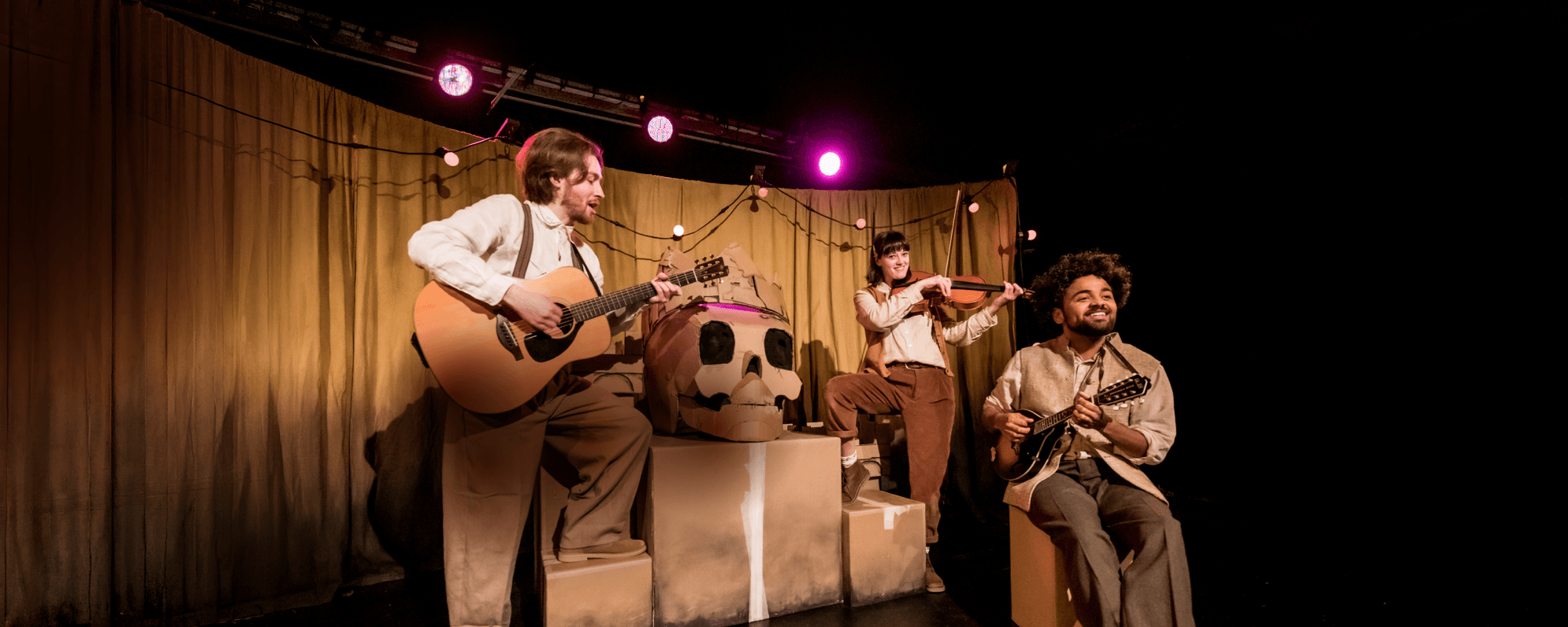 Three musicians, wearing brown and beige, play two guitars and a violin as they gather around cardboard boxes. On top of the boxes sits a giant cardboard skull with a crown.