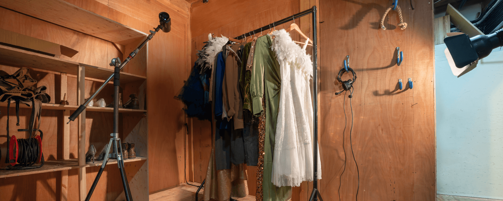 In a wooden room, clothes hang on a rail beside a black spotlight. Headphones are also hanging on one of the walls.