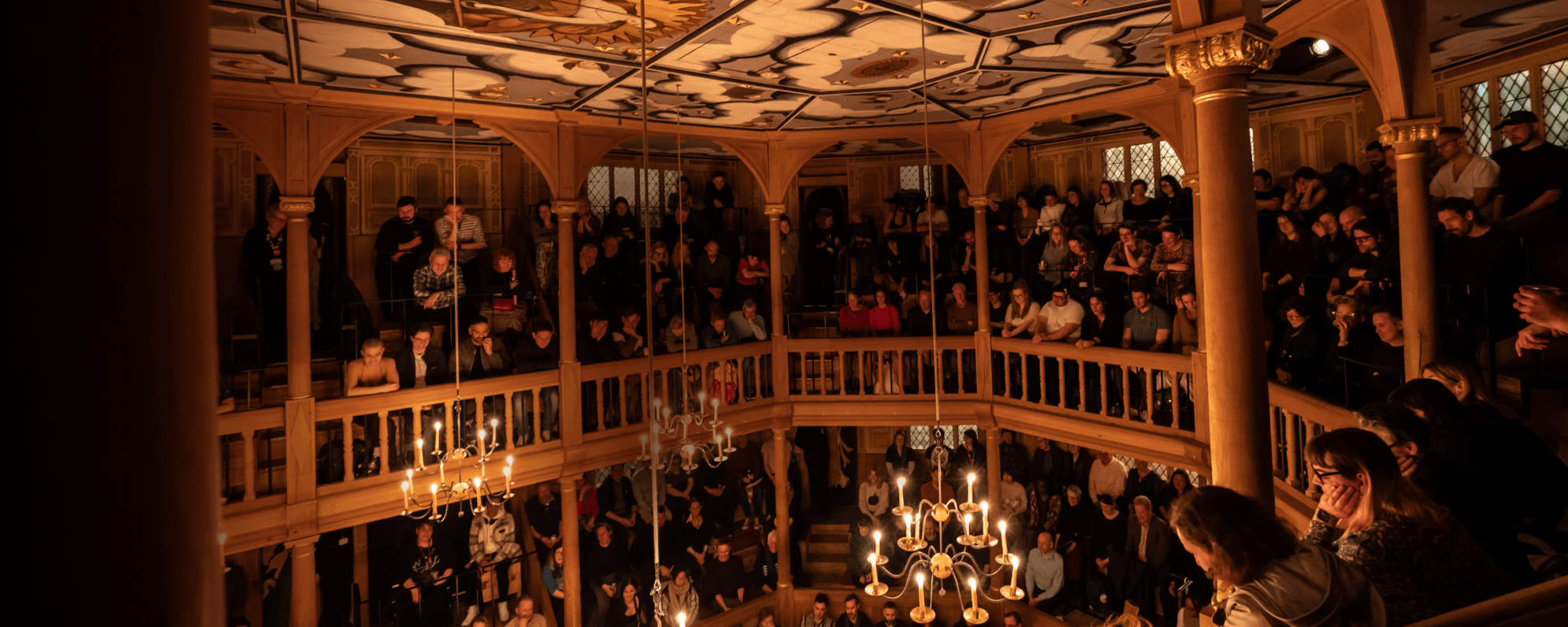 The interior of a candlelit wooden playhouse filled with a seated audience.
