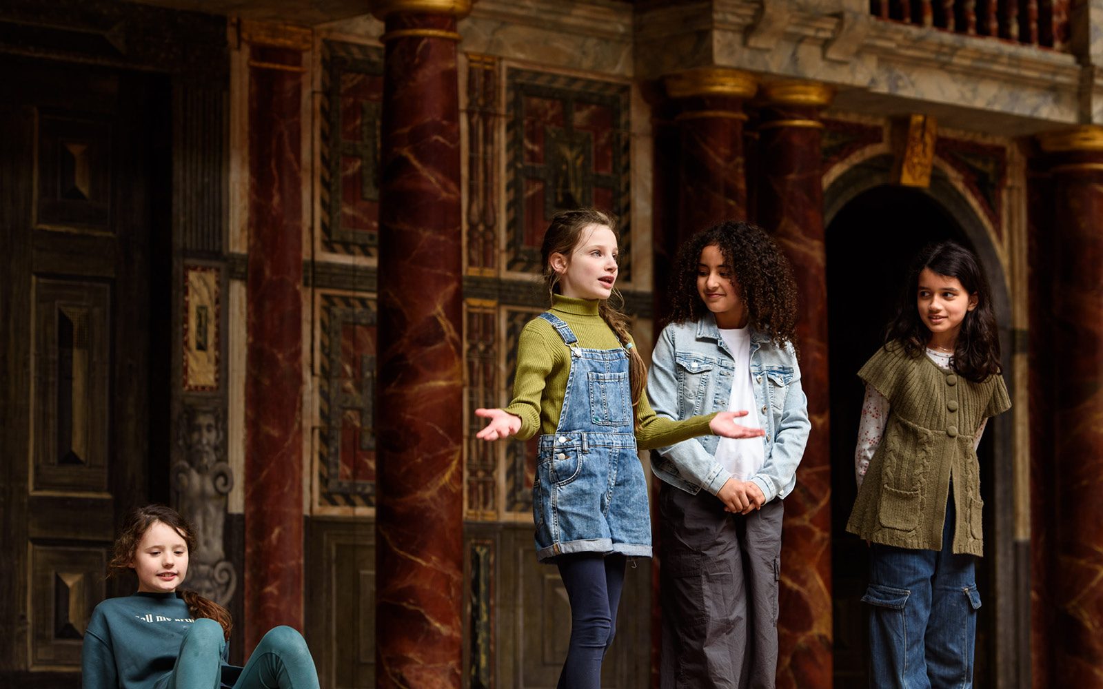 Three children stand in a line. One has their hands outstretched and the others are looking at them. Another child sits on the floor nearby. In the background are red marble pillars and wooden doors.