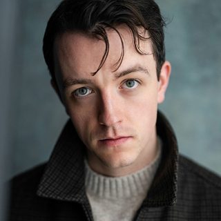 Jordan Rhys wearing a dark collared jacket and looking directly at the camera.