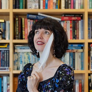Kaz Penney holding a quill in front of a bookcase.
