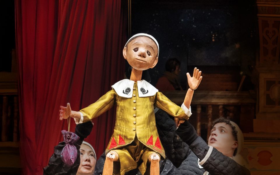 Two people hold a wooden puppet above their heads. The puppet wears a gold jacket with a white collar and is waving one hand in the air.