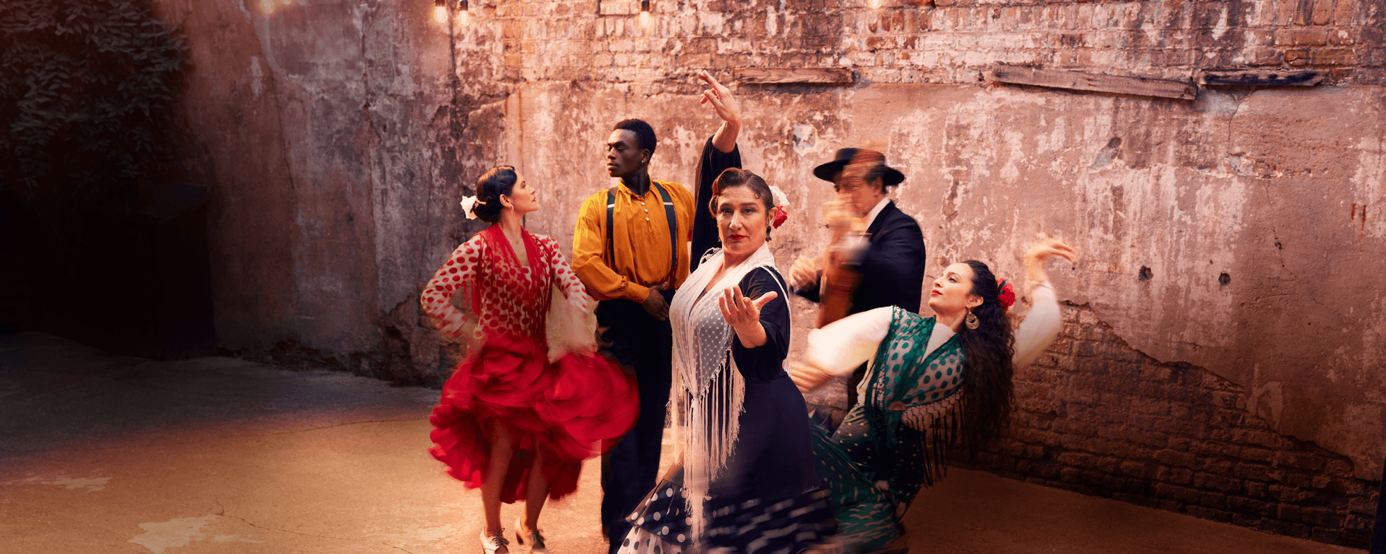 A group of flamenco performers are captured in motion against a plain brick wall. Four of the group, wearing traditional colourful flamenco costumes, take strong dance positions. A fifth person wearing a dark suit stands playing a guitar, which is blurred through the capture of movement.