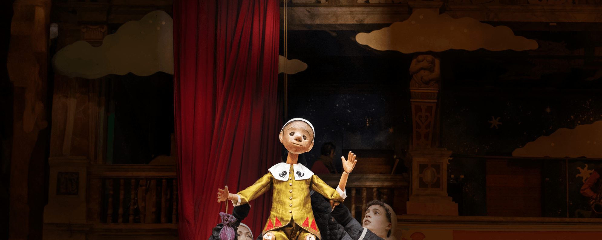 Two people hold a wooden puppet above their heads. The puppet wears a gold jacket with a white collar and is waving one hand in the air.