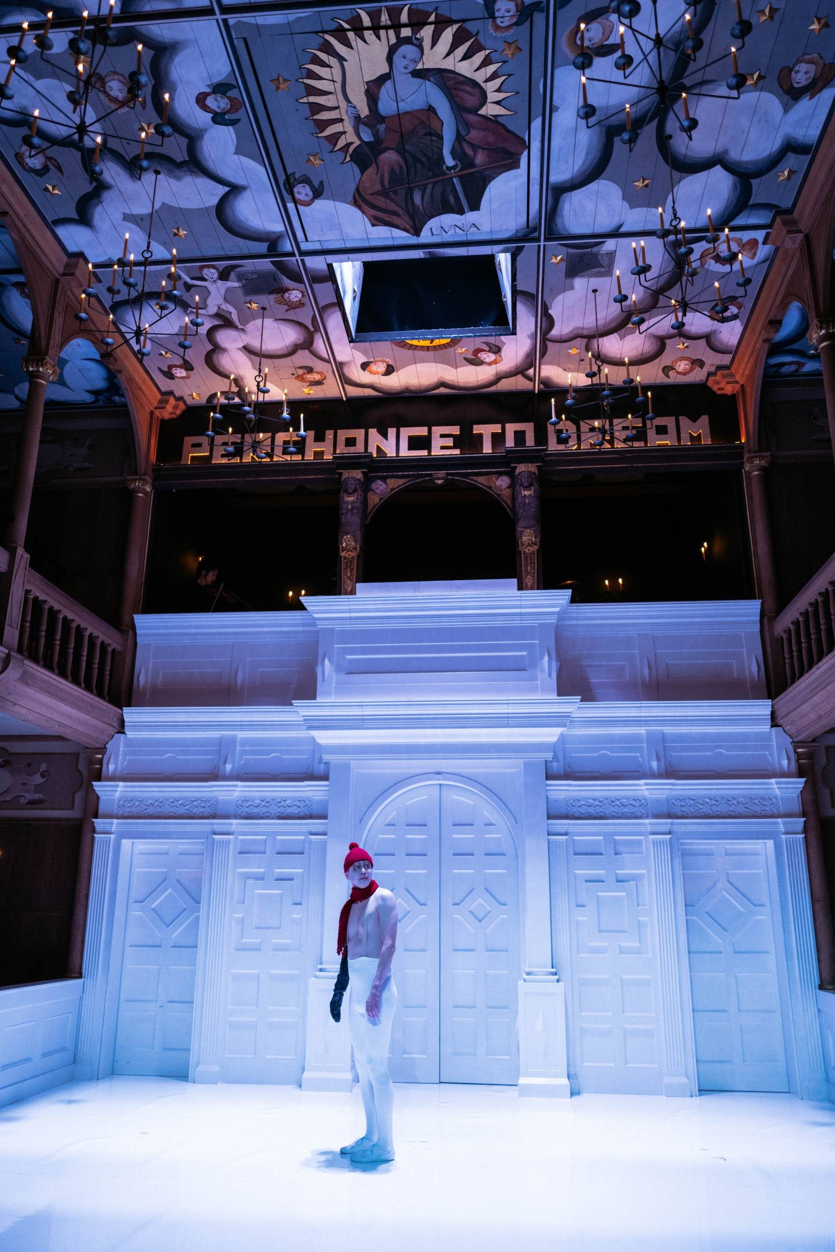 An actor wearing a red bobble hat and scarf and white ballet tights stands in the middle of a stage with white floors, white walls, and a shadowy balcony area. The ceiling is covered in medieval-style painting of angels, mythological figures and clouds. Gold block letters above the balcony read 'Perchance to dream'.