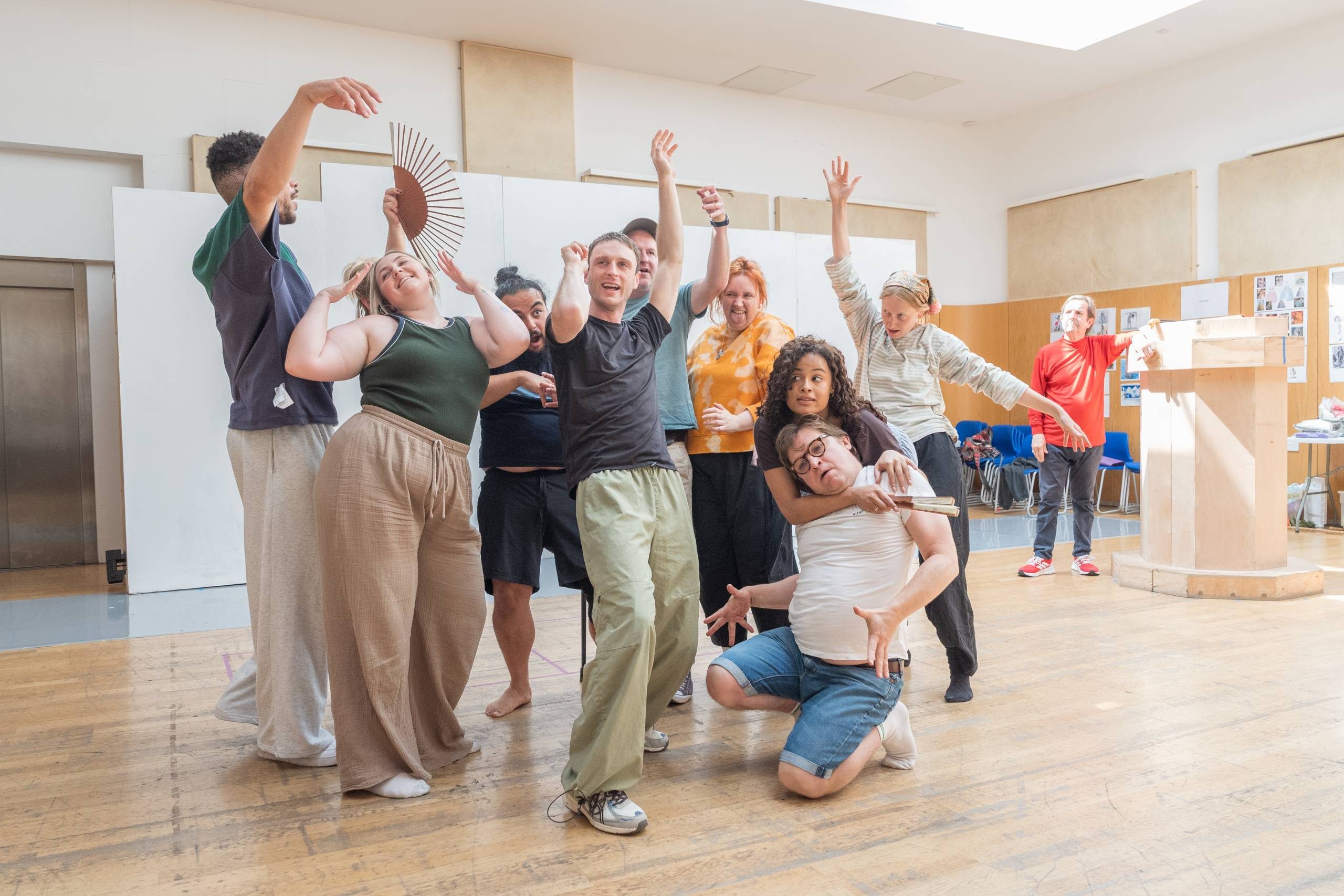 A diverse group of people raise their arms and dance in a wooden rehearsal room. One person holds a fan, and one person wearing a red top, leaning on a wooden pillar, watches from the side.