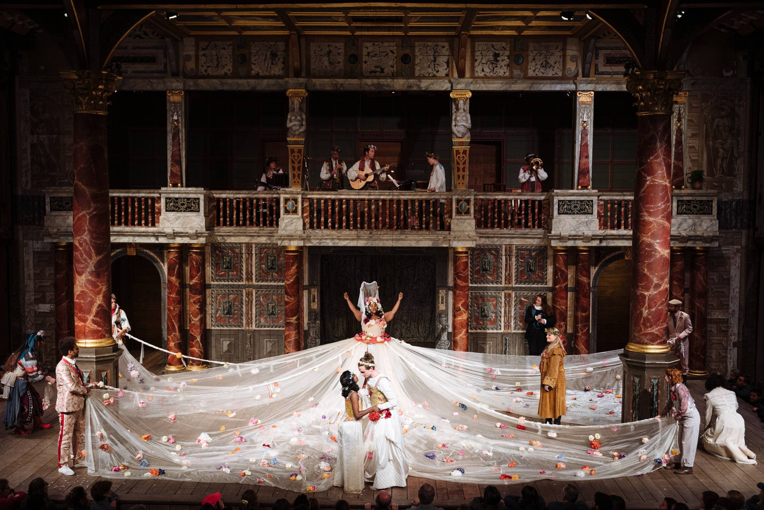 A tableau of the Globe Theatre which is comprised of a wooden stage and red marbled pillars. An actor suspended on wires in the centre of the stage wears a white dress with large flowers that encompasses the stage. People hold the skirt and a couple at the front of the stage hold hands. There is a band of musicians on a balcony above the stage.