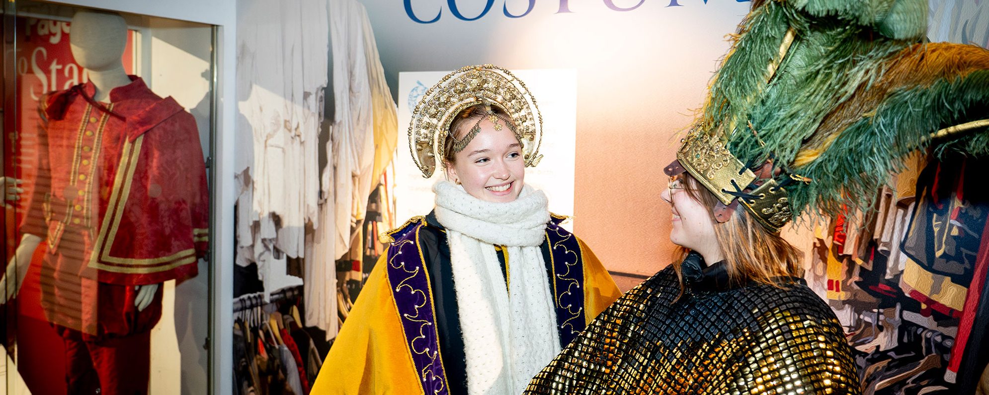 Two people wearing costumes smile at each other. One wears a jewelled headdress and the other wears a a hat with green feathers. Around them, other costumes are displayed or hanging up.