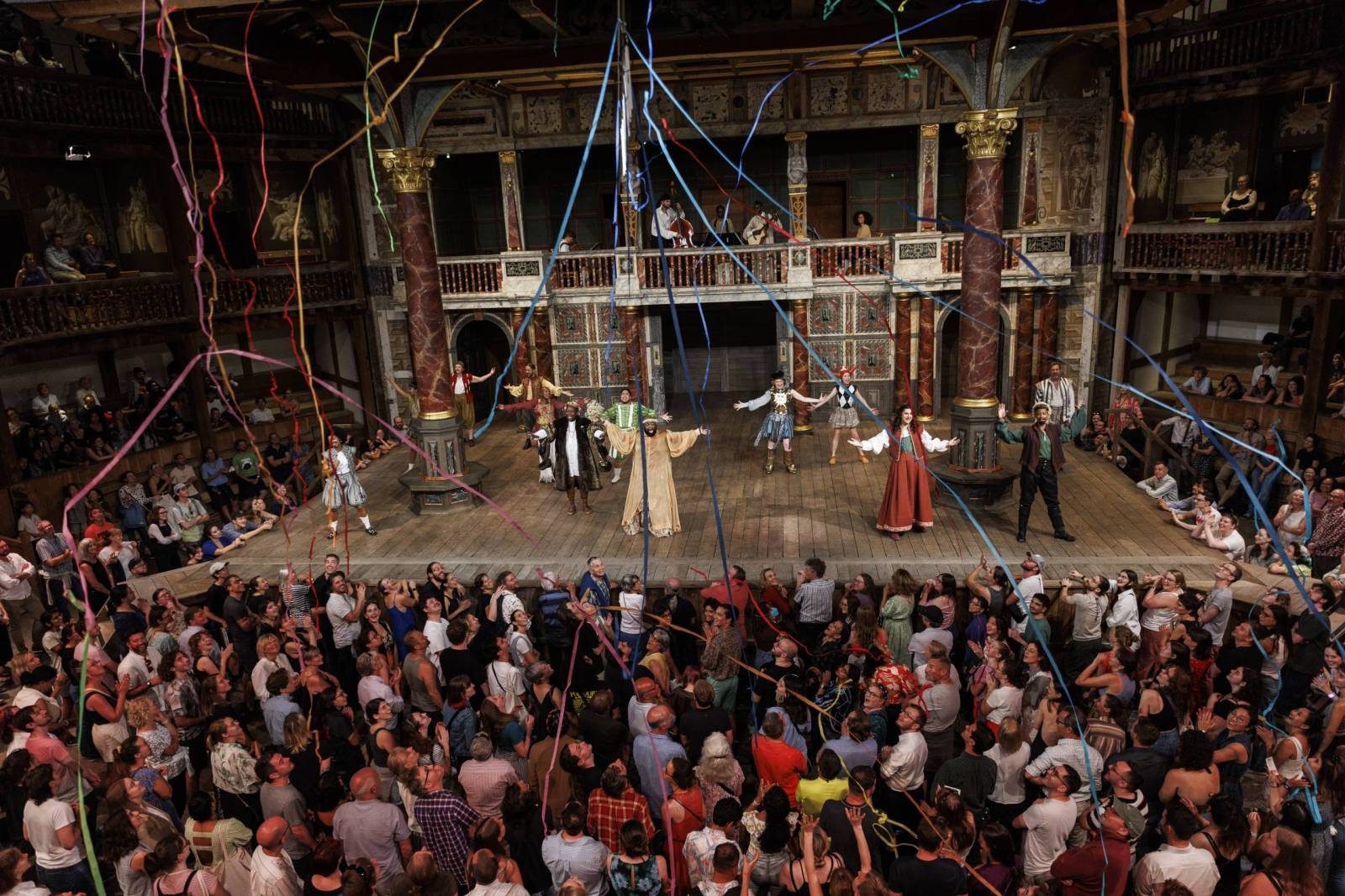 A group of actors stand on the wooden Globe Theatre stage with their arms held wide facing a packed audience. Multi-coloured confetti streamers fall from above.