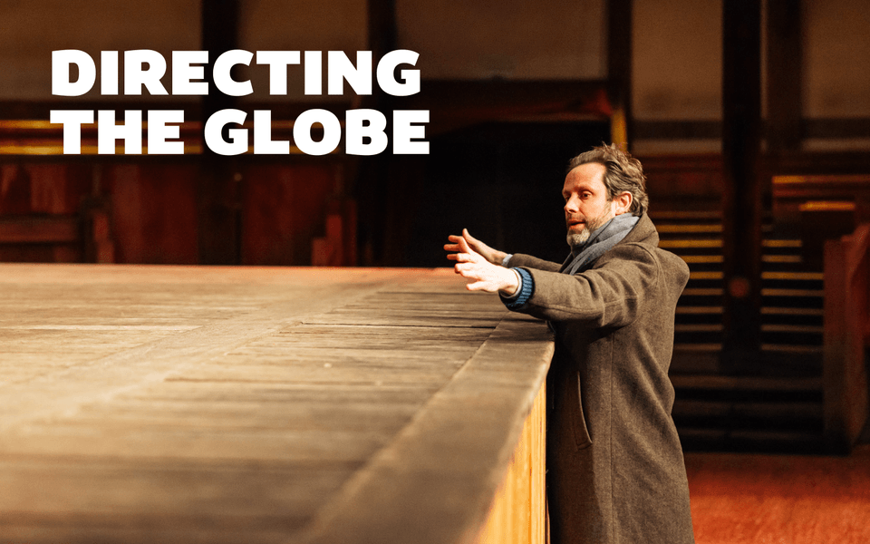Directing the Globe. A person standing in front of wooden stage, holding their arms out in front of them.
